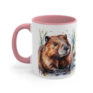 Könnte beinhalten: Eine weiße Keramiktasse mit rosafarbenem Rand zeigt eine Aquarell-Illustration eines braunen Bibers mit buschigem Schwanz, der im Wasser mit grünen Schilf und blauen und weißen Aquarellspritzer im Hintergrund sitzt.