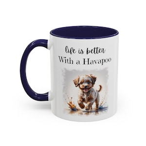 May include: A white ceramic mug with a dark blue handle and interior rim. The front features a graphic of a light brown Havapoo puppy splashing in water, with the black text "life is better With a Havapoo" printed above the puppy illustration.