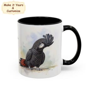 Puede incluir: Una taza de cerámica blanca con asa y borde interior negros, que presenta una ilustración en acuarela de una cacatúa negra con plumas rojas en la cola. Sobre la taza, una pequeña etiqueta beige muestra el texto Make it Yours & Customize.