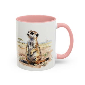 Könnte beinhalten: Ein weißer Keramikbecher mit einem zartrosa Innenraum und Henkel. Die Außenseite zeigt eine detaillierte Aquarellillustration eines Erdmännchens, das aufrecht in einer Wüstenlandschaft mit spärlicher Vegetation und Horizont steht.