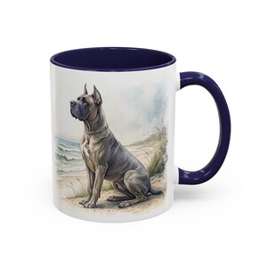 May include: A white ceramic mug with a dark blue interior and handle, featuring a watercolour illustration of a fawn-coloured Cane Corso dog sitting on a sandy beach with ocean waves and tall grass in the background, rendered in soft tones.