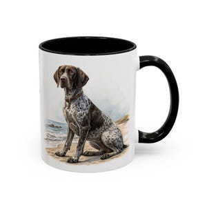 May include: White ceramic mug with a black interior and handle. The mug features a watercolour illustration of a German Shorthaired Pointer dog on a beach. The dog is brown and white with a brown collar.