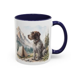 May include: White ceramic mug with a navy blue interior and handle. The mug features a watercolour illustration of a dog sitting near a tent in a mountain landscape. The dog is brown and grey with a brown collar.
