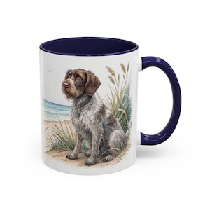 Könnte beinhalten: Weißer Keramikbecher mit marineblauem Innenraum und Griff. Der Becher zeigt eine Aquarellillustration eines Hundes, der an einem Strand mit hohem Gras und dem Ozean im Hintergrund sitzt. Der Hund ist braun und grau.