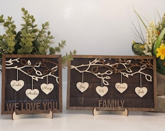 May include: Two wooden signs with a tree branch design and hearts hanging from the branches. The hearts have names written on them. The first sign says "We Love You" and the second sign says "Family".