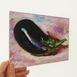 May include: An original oil painting of an eggplant. The artwork features a dark purple eggplant with a green stem against a background of pink, purple, and blue brushstrokes. The painting is held by a hand.