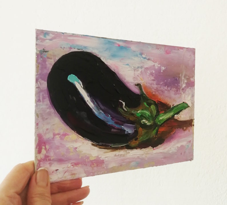 May include: An original oil painting of an eggplant. The artwork features a dark purple eggplant with a green stem against a background of pink, purple, and blue brushstrokes. The painting is held by a hand.