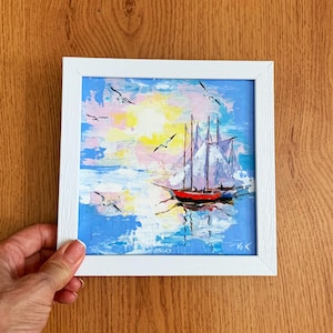 May include: A framed painting of a sailboat with white sails on a blue ocean with a pink and yellow sky. The painting is in a white frame.