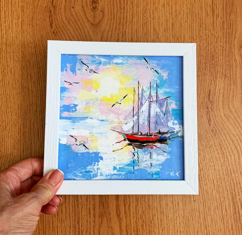May include: A framed painting of a sailboat with white sails on a blue ocean with a pink and yellow sky. The painting is in a white frame.