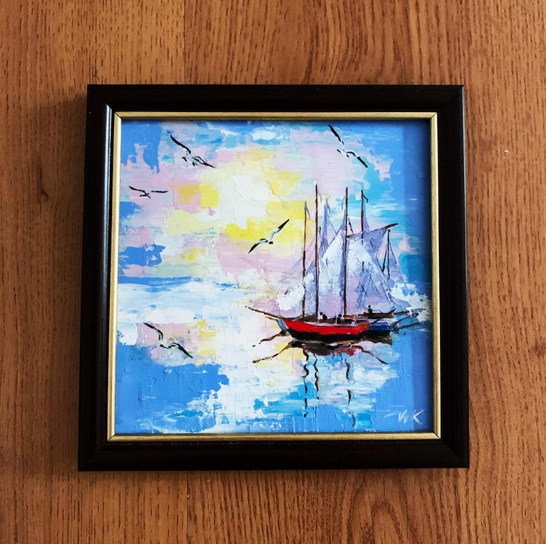 May include: A framed painting of a sailboat with white sails on a blue sea with white clouds and a yellow sun. The painting is in a black frame with a gold inner border.