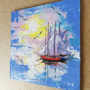 May include: A painting of a sailboat with white sails on a blue and pink water background with a yellow sun and white birds flying in the sky.