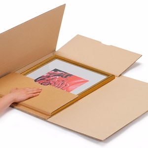 May include: A framed picture with a red rose is being carefully placed into a cardboard box with a protective layer of cardboard.