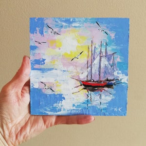 May include: A painting of a sailboat with white sails on a blue body of water with a pink and yellow sky. The sailboat has a red hull and is surrounded by white clouds. There are several seagulls flying in the sky.