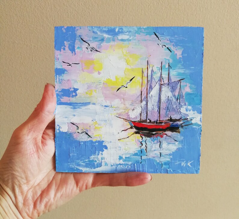 May include: A painting of a sailboat with white sails on a blue body of water with a pink and yellow sky. The sailboat has a red hull and is surrounded by white clouds. There are several seagulls flying in the sky.