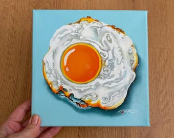 May include: A painting of a fried egg on a light blue background. The egg yolk is bright orange and the white is white with a slightly brown edge.