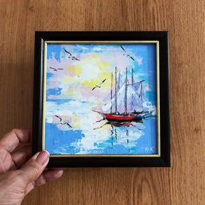May include: A framed painting of a sailboat with white sails on a blue and pink seascape with white birds flying overhead.