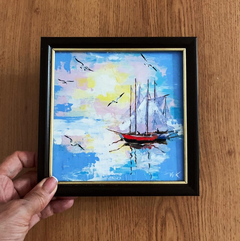 May include: A framed painting of a sailboat with white sails on a blue and pink seascape with white birds flying overhead.