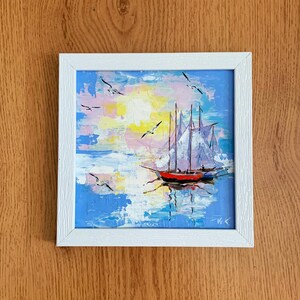 May include: A framed painting of a sailboat with white sails on a blue and white seascape with a yellow sun and pink clouds. The painting is in a white frame.