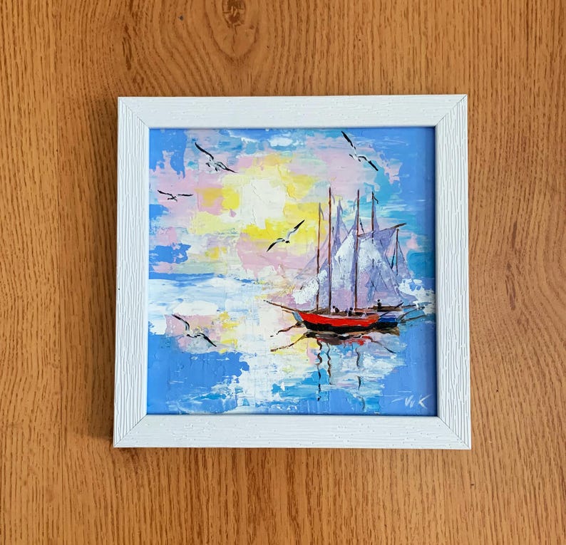 May include: A framed painting of a sailboat with white sails on a blue and white seascape with a yellow sun and pink clouds. The painting is in a white frame.
