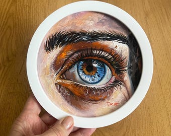 Puede incluir: Pintura al óleo redonda enmarcada en blanco, que representa un ojo humano. El ojo es de un azul vibrante con un iris marrón y largas pestañas. La piel alrededor del ojo está pintada en tonos naranja, rosa y marrón, con cejas oscuras.