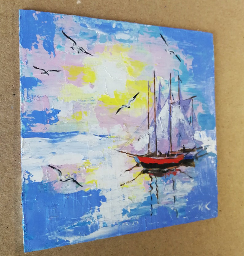 May include: A painting of a sailboat with white sails on a blue and pink sky with white clouds. The sailboat is red and black and is reflected in the water. There are seagulls flying in the sky.