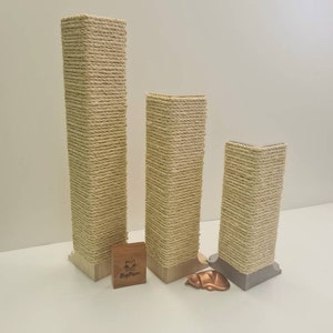 May include: Three beige cat scratching posts of varying heights, wrapped in natural sisal rope. The posts are made of wood and have a square base. The smallest post is on the right, the middle post is in the centre, and the tallest post is on the left. The posts are on a white surface. The brand name "BigPaw" is visible on a small wooden block in front of the posts.