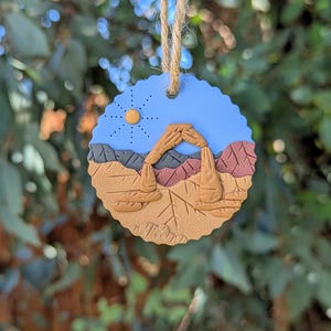 May include: A round ornament with a blue sky background and a sun design. The ornament features a detailed landscape with a brown arch, brown and red rock formations, and a scalloped edge. A natural twine loop is attached for hanging.