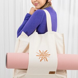 May include: A cream-colored canvas tote bag featuring an embroidered orange sun design on the front. The bag has a horizontal strap across the center designed to hold a rolled-up light pink yoga mat securely in place. A small brown label is attached.