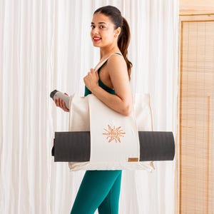 May include: A cream-coloured canvas tote bag featuring an orange embroidered sun design on the front. The bag has a wide horizontal strap across the centre designed to hold a rolled-up dark grey yoga mat securely in place while being carried.