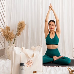 May include: A cream-coloured canvas tote bag featuring a brown sun graphic on the front and a small rectangular brand tag. Next to the bag stands a light grey insulated stainless steel water bottle with a black lid and a visible brand logo.