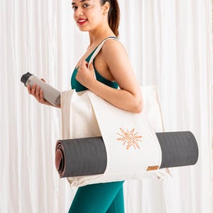 May include: A cream-coloured canvas tote bag featuring a brown embroidered sun design, designed to carry a rolled dark grey yoga mat. The bag includes a small brown rectangular brand tag on the lower right side and has two shoulder straps.