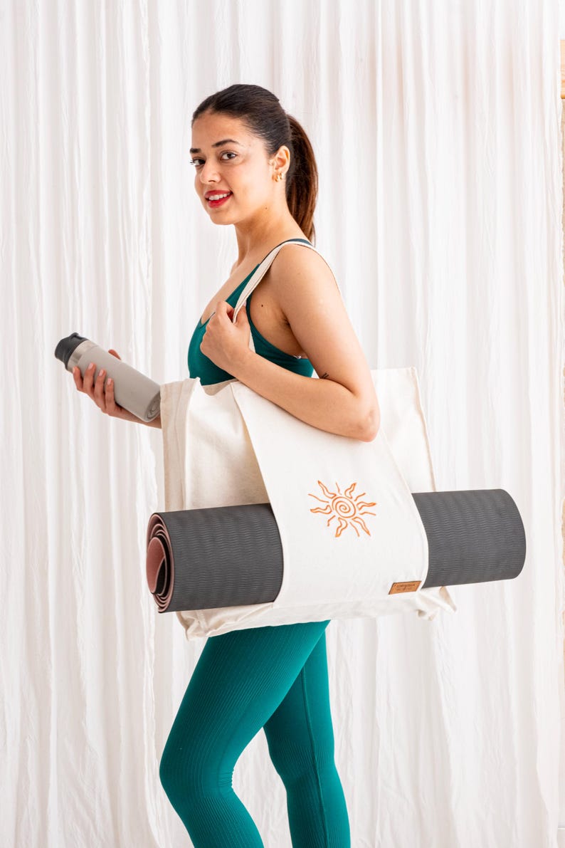 May include: A cream-coloured canvas tote bag featuring a brown embroidered sun design, designed to carry a rolled dark grey yoga mat. The bag includes a small brown rectangular brand tag on the lower right side and has two shoulder straps.