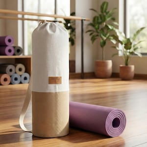 May include: A cylindrical yoga mat bag featuring a two-tone design with an off-white upper section and a tan lower section. A brown rectangular label with the text LORENZELLA is centred on the bag. A purple rolled yoga mat lies horizontally nearby.