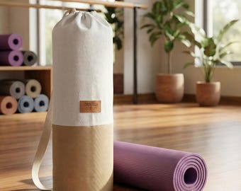 Könnte beinhalten: Eine zylindrische Yogamatten-Tasche im zweifarbigen Design mit einem cremeweißen oberen Teil und einem beigen unteren Teil. Ein braunes rechteckiges Etikett mit dem Text LORENZELLA ist mittig auf der Tasche angebracht. Eine violette Yogamatte liegt daneben.