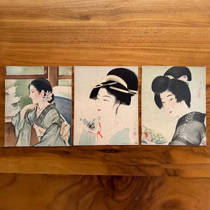 May include: Three vintage Japanese postcards featuring illustrations of women in traditional clothing. The postcards are printed on cream-colored paper with a light brown border. The women are depicted with delicate features and intricate hairstyles. The postcards are in excellent condition.