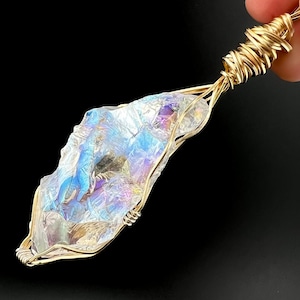 Puede incluir: Un colgante de cristal envuelto en alambre dorado. El cristal iridiscente tiene una forma natural e irregular y muestra una gama de colores, incluyendo azules, morados y verdes. El colgante es sostenido por una mano sobre un fondo negro.