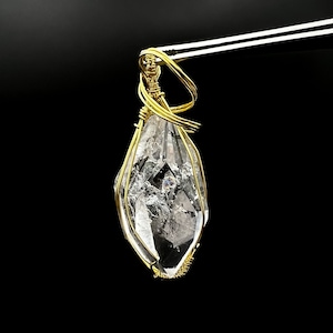 Puede incluir: Un colgante de cristal de cuarzo transparente envuelto en alambre dorado. El cristal está facetado y tiene forma de lágrima.