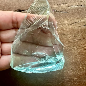 May include: A triangular piece of translucent, pale blue glass with a smooth, polished surface and visible internal fracture lines. The glass has a sharp, pointed top edge and a wider, slightly curved base, displaying a clear, icy appearance.