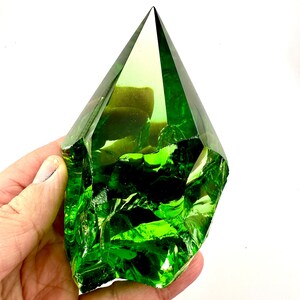 May include: A large, green, faceted glass crystal in the shape of a pyramid. The crystal is clear and has a slight yellow tint. It is being held in a hand.