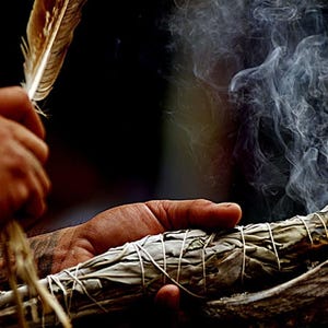 May include: A person holding a feather and a bundle of burning sage, with smoke rising. The sage is wrapped with twine and held in a shell. The background is dark, emphasizing the smoke and the objects.