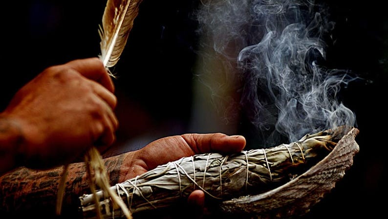 May include: A person holding a feather and a bundle of burning sage, with smoke rising. The sage is wrapped with twine and held in a shell. The background is dark, emphasizing the smoke and the objects.