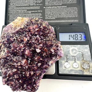 May include: A purple amethyst crystal cluster weighing 148.3 grams on a digital scale. The scale has a black display screen with white numbers and a black background. The crystal is a beautiful shade of purple and has a variety of shapes and sizes.