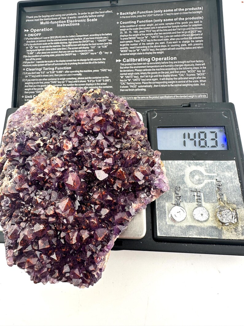 May include: A purple amethyst crystal cluster weighing 148.3 grams on a digital scale. The scale has a black display screen with white numbers and a black background. The crystal is a beautiful shade of purple and has a variety of shapes and sizes.