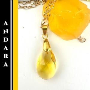 May include: A gold-colored necklace with a teardrop-shaped, yellow pendant. The pendant is faceted and has a gold-colored cap. The necklace chain is also gold-colored. The word "ANDARA" is printed vertically on the left side of the image.