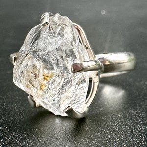 Puede incluir: Un anillo de cristal en bruto transparente con una banda de plata. El cristal en bruto tiene una forma irregular y está montado sobre una banda de plata pulida. El cristal tiene un aspecto ligeramente translúcido.