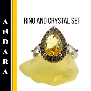 Puede incluir: Un anillo plateado con una gran gema amarilla en forma de pera, rodeada de piedras claras más pequeñas. El anillo está sobre un cristal amarillo. La imagen incluye el texto "RING AND CRYSTAL SET".