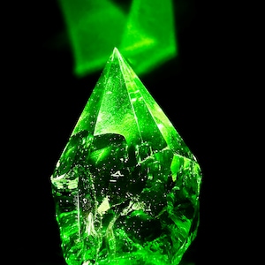 May include: A large, faceted, green crystal with a pointed top. The crystal is illuminated from above, creating a bright green glow.