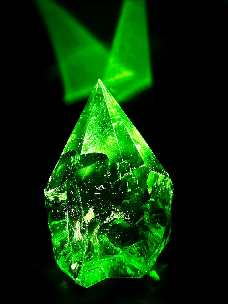 May include: A large, faceted, green crystal with a pointed top. The crystal is illuminated from above, creating a bright green glow.