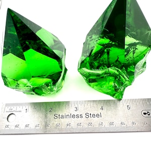 May include: Two green glass pyramid-shaped crystals, each with a different shape and texture, are displayed on a white surface with a stainless steel ruler for scale.