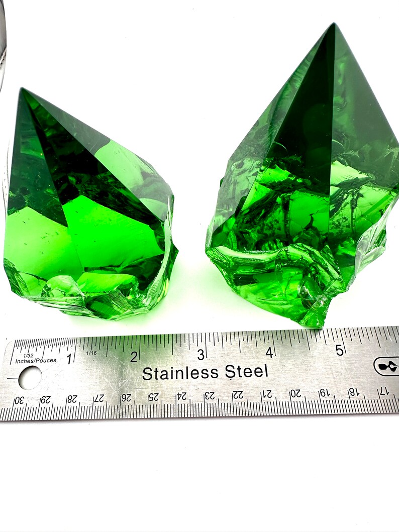 May include: Two green glass pyramid-shaped crystals, each with a different shape and texture, are displayed on a white surface with a stainless steel ruler for scale.